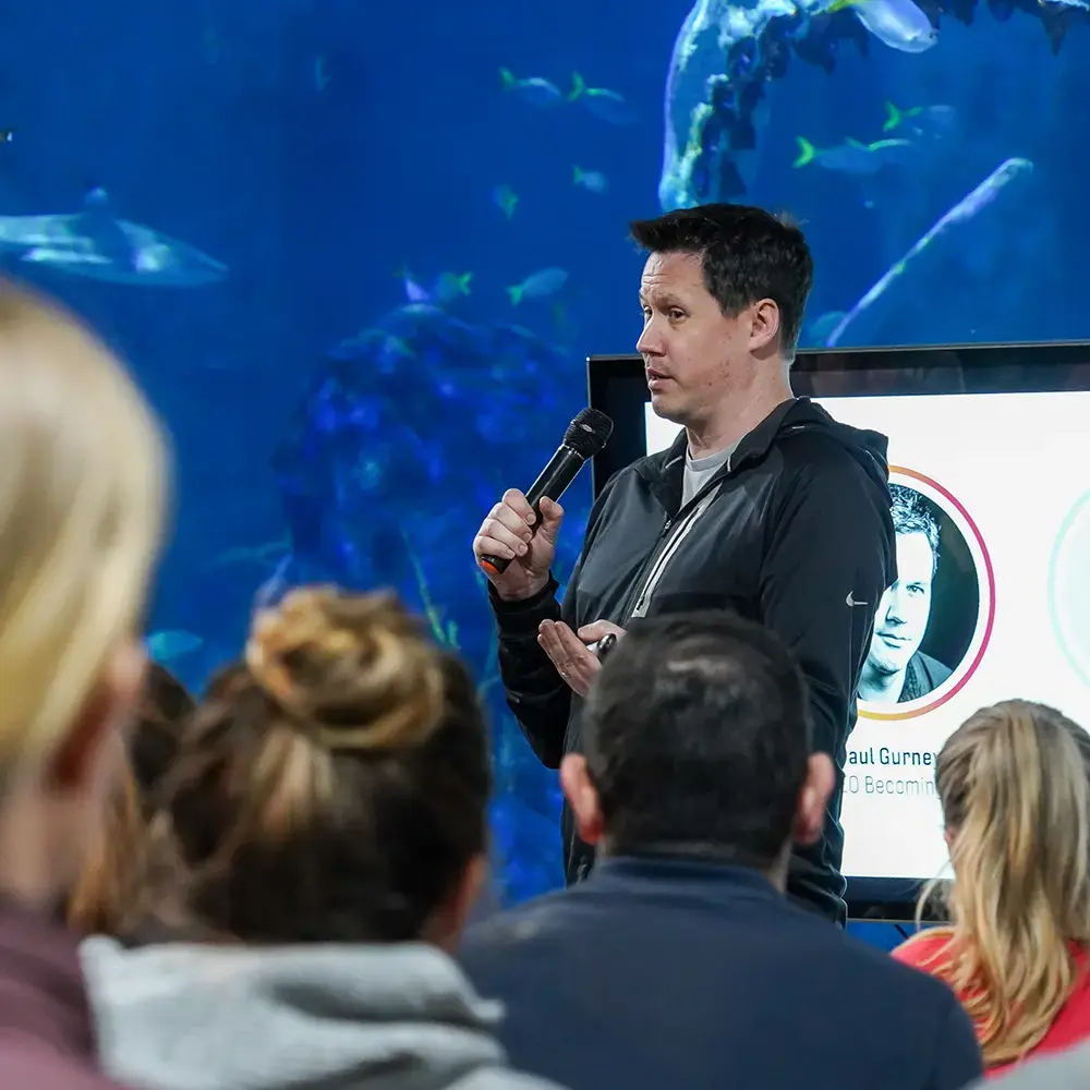 Paul Gurney, CEO of BecomingX, speaking to the Commonwealth Games team in front of an aquarium backdrop.