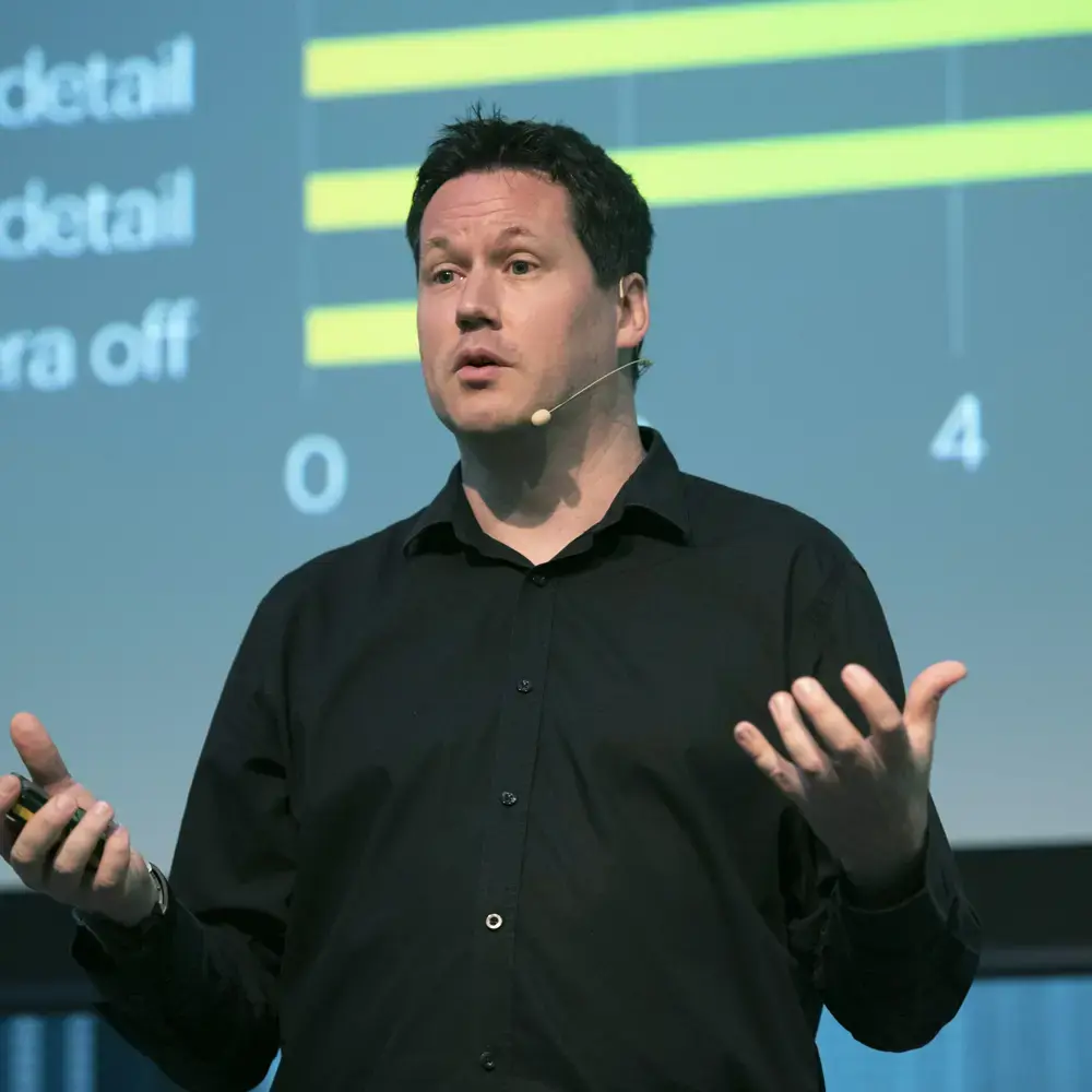 Paul Gurney presenting on stage at an Accenture event, with a data chart displayed behind him.
